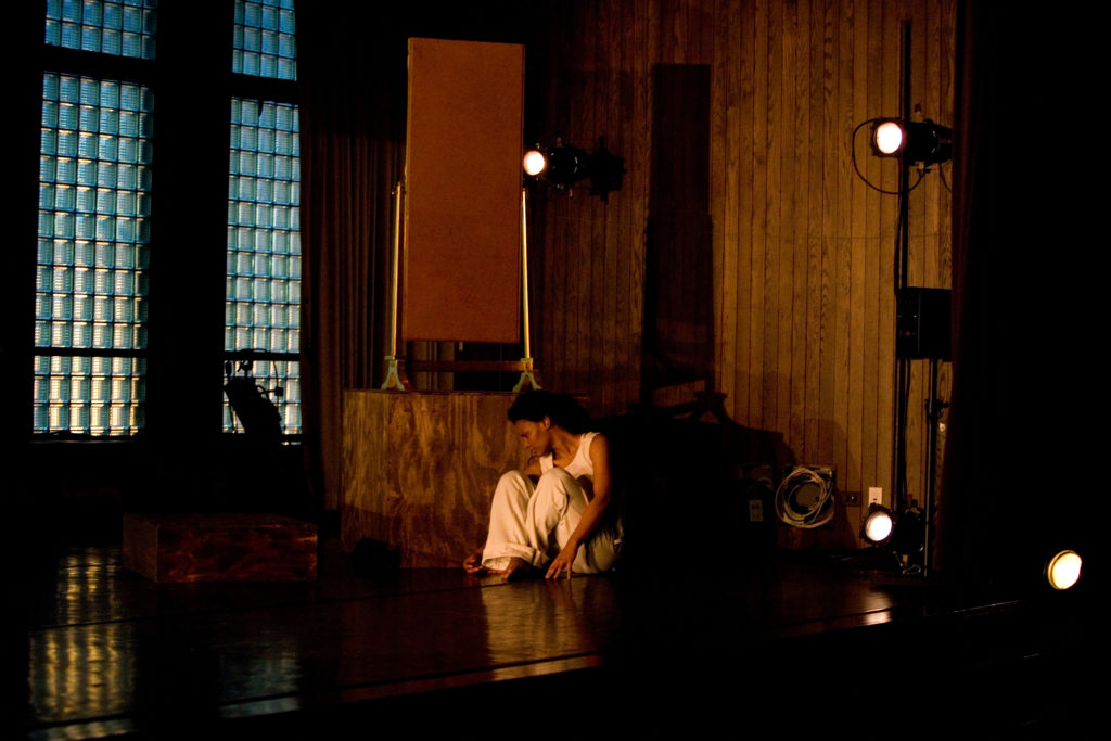 A still photo of a woman wearing a white outfit, sitting on the ground in the middle of a stage looking at the ground. There are camera lights to the side.