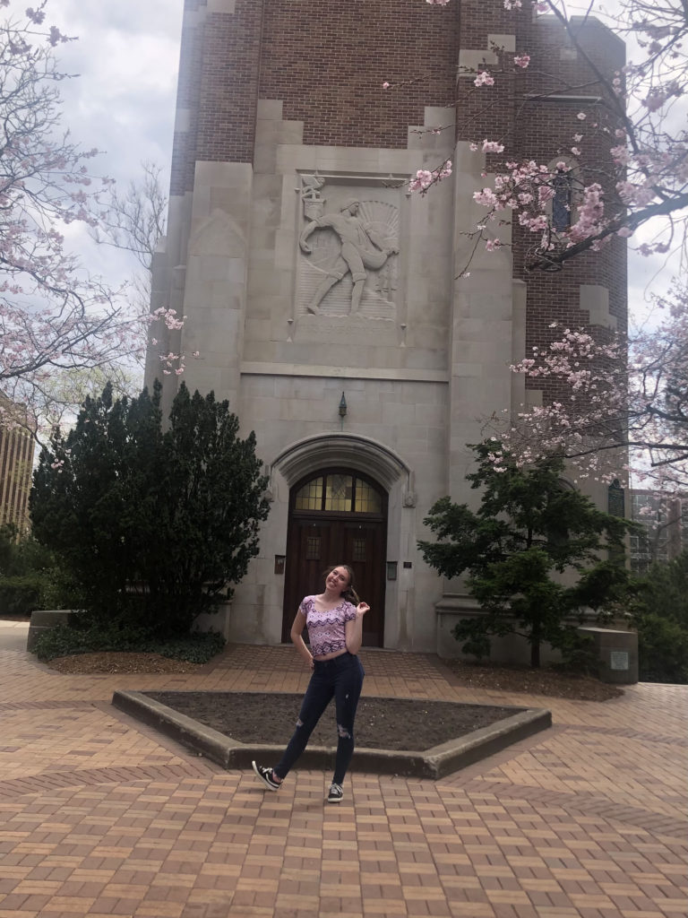 Girl in pink shirt poses in front of a tower and trees.