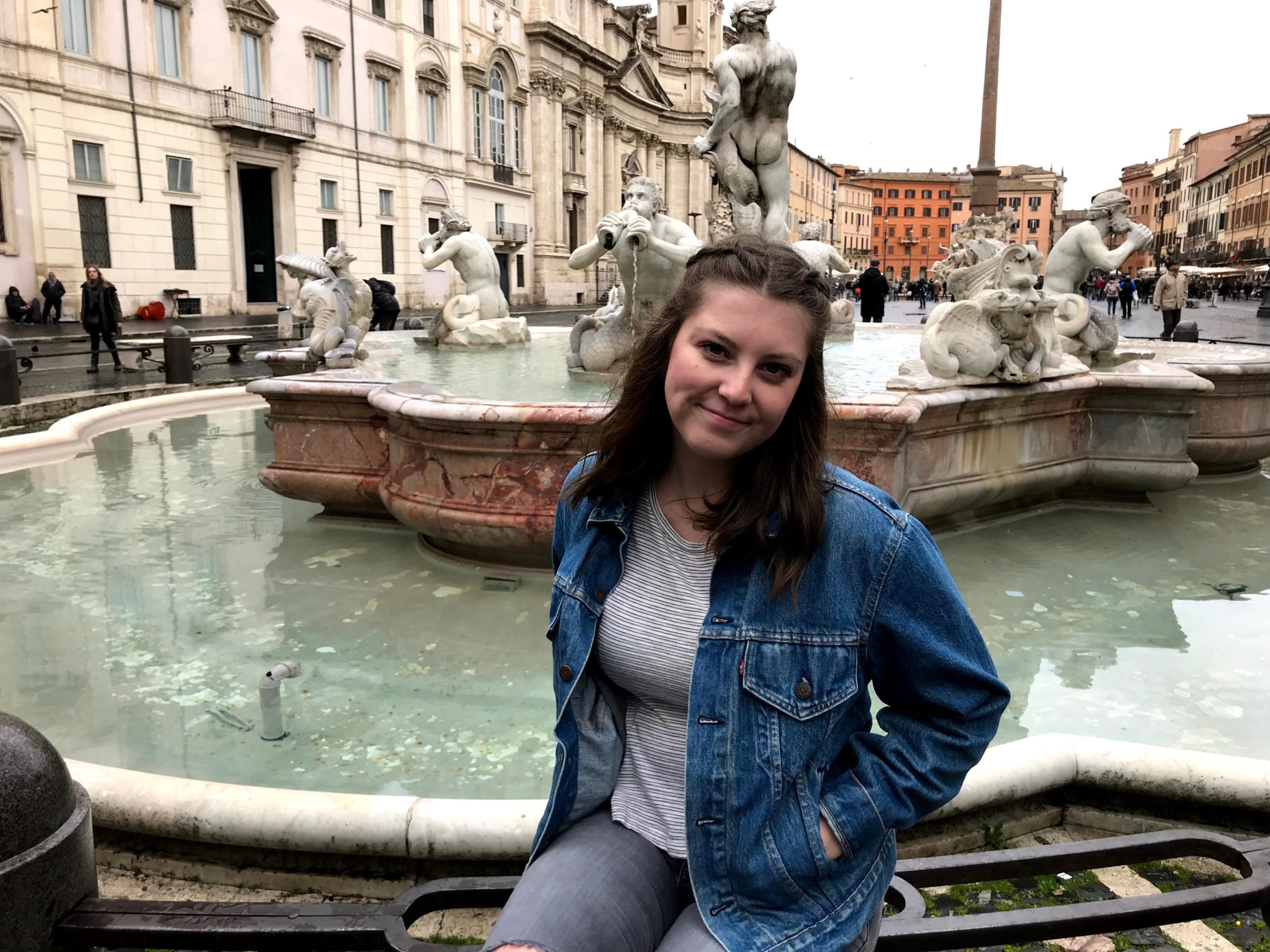 A young woman sitting in front of a fountain with buildings in the background.