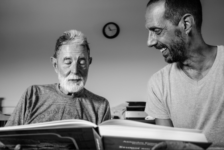 Two men sitting side by side reading from the same book.