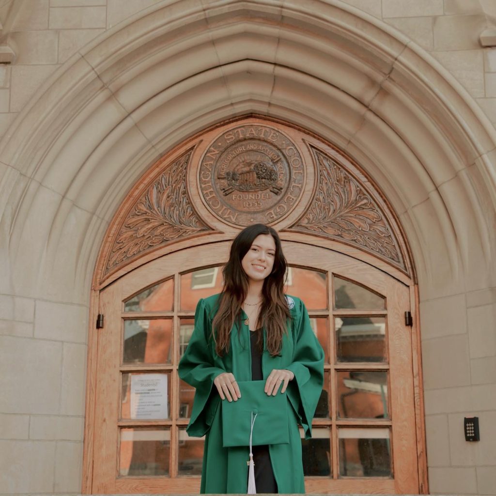 Woman with long brown hair is standing in front of a doorway and wearing a green graduation gown. She is holding a green graduation cap in front of her torso.