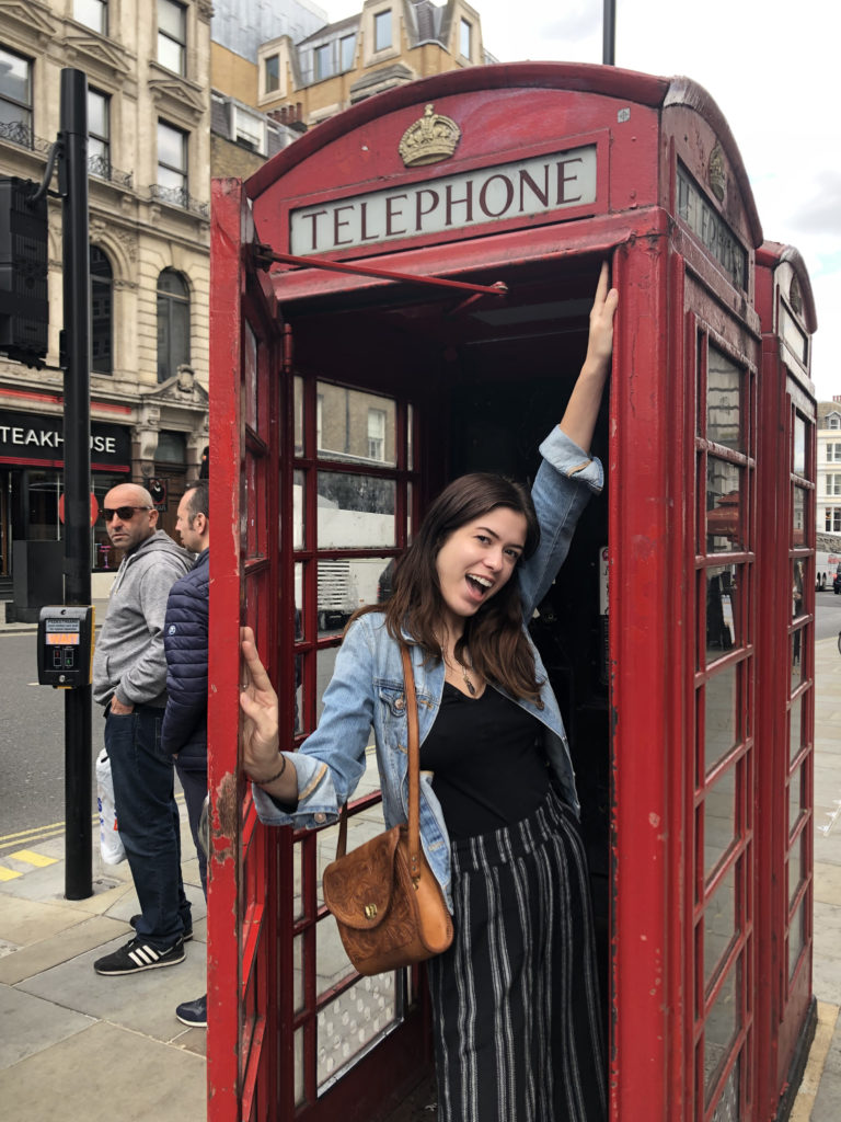 Woman with short brown hair is wearing a denim coat and black and white striped pants. She is standing in side of a red telephone booth in England.