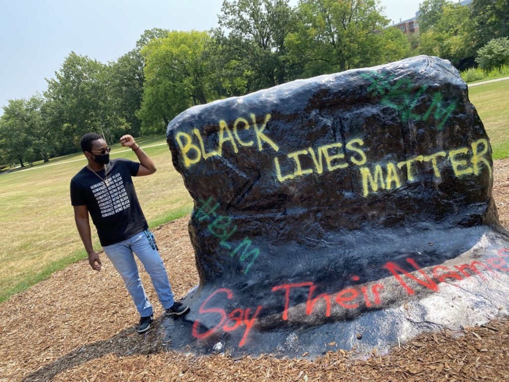 A young man stands by a large boulder with the words 'BLACK LIVES MATTER' written in yellow spray paint.