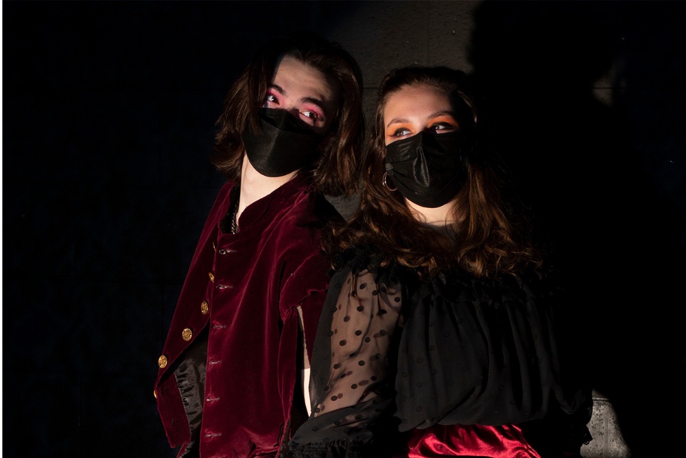 Two people stand back to back, both with brown hair and wearing black face masks.