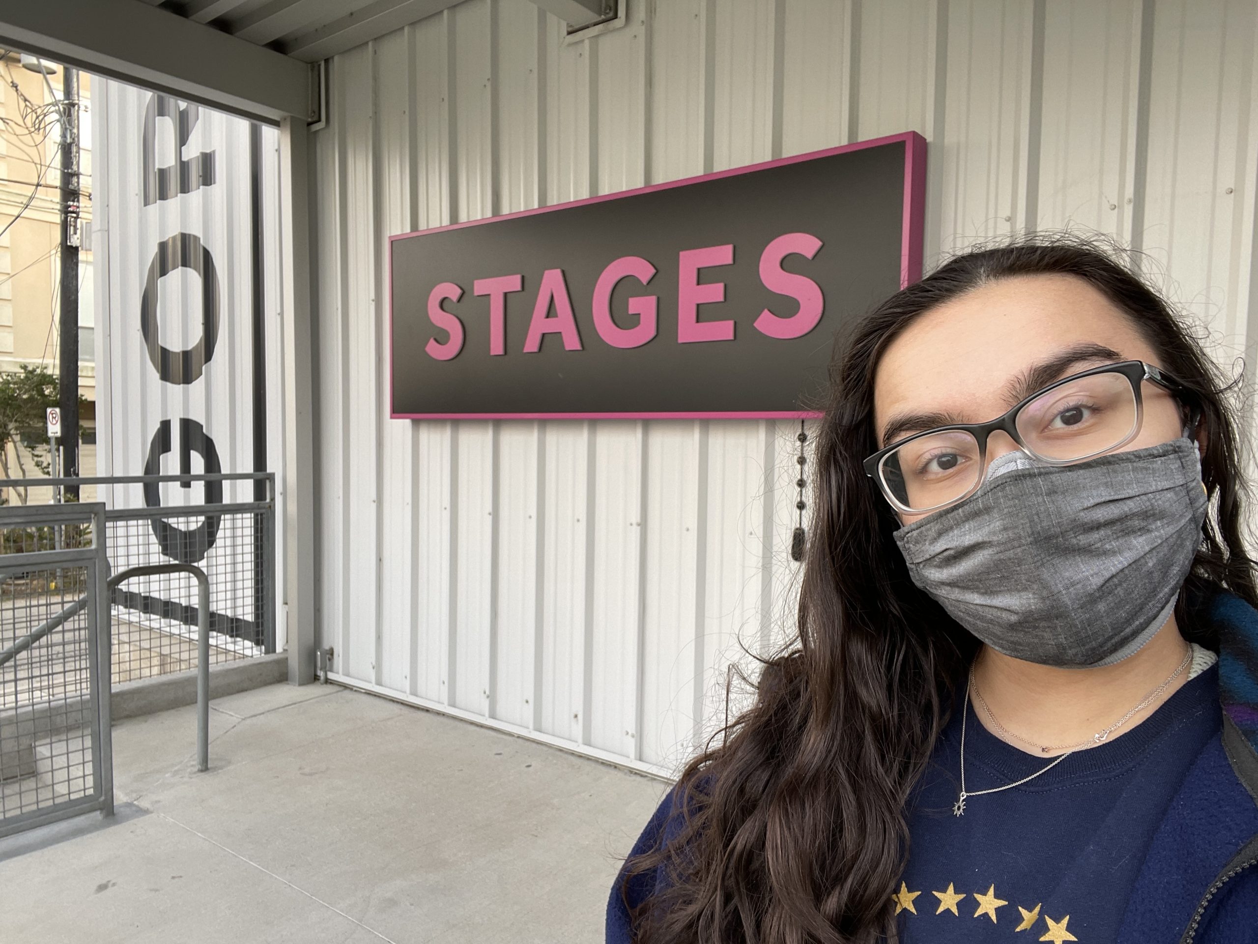 Woman is wearing glasses and a face mask and standing in front of a sign that says "Stages"
