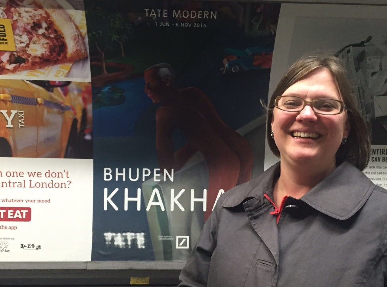 Woman wearing glasses and a black trenchcoat is standing in front of an advertisement for a museum exhibition. 