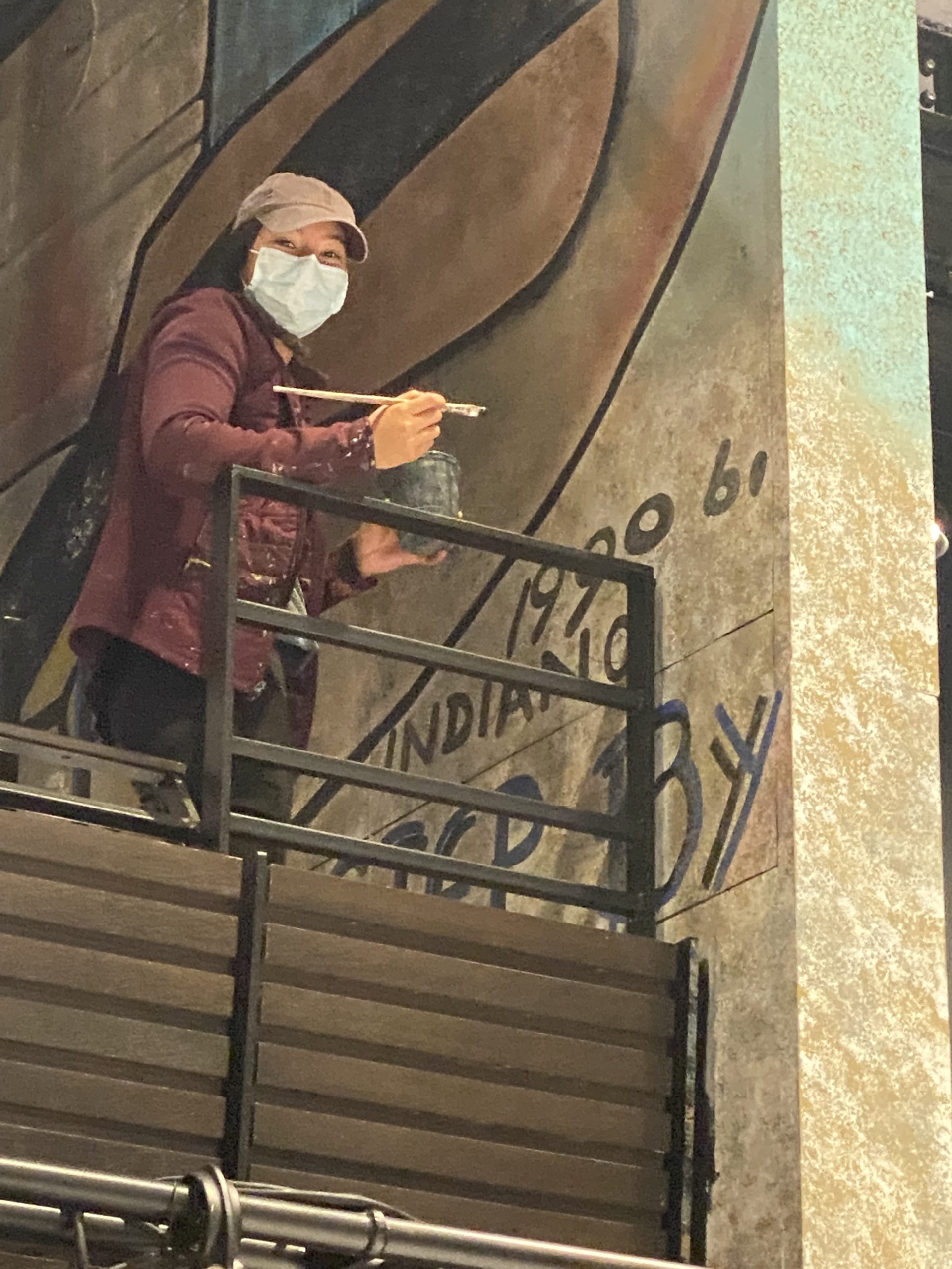 Woman wearing a face mask and a red jacket is holding a paintbrush and is standing next to a wall she is painting.