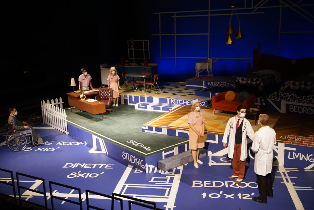 A wide angle photo of a stage that looks like blueprints. Actors are posed across the stage, dressed as nurses and doctors.