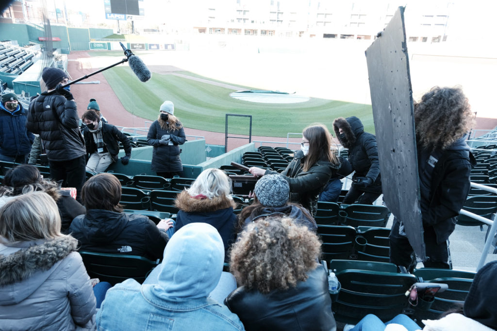 Filmmakers with audio and film equipment attempt another take of extras sitting in the stands of a baseball stadium.