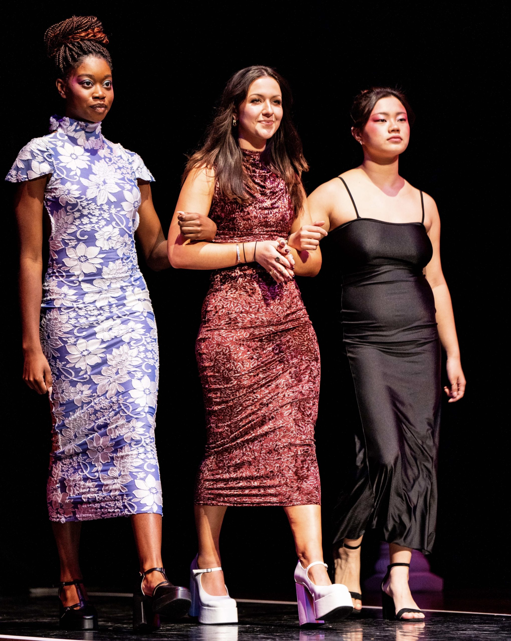 Three young women in intricate dresses walk down the stage arm in arm with each another.