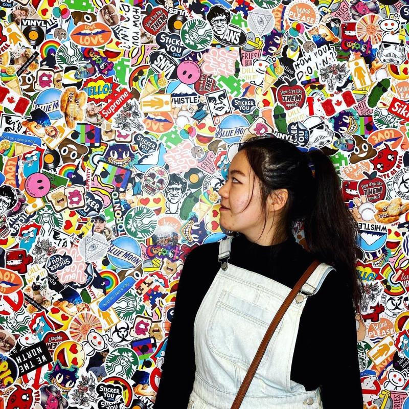 Woman with black hair wearing overalls and a black shirt is standing in front of a wall with numerous stickers on it.