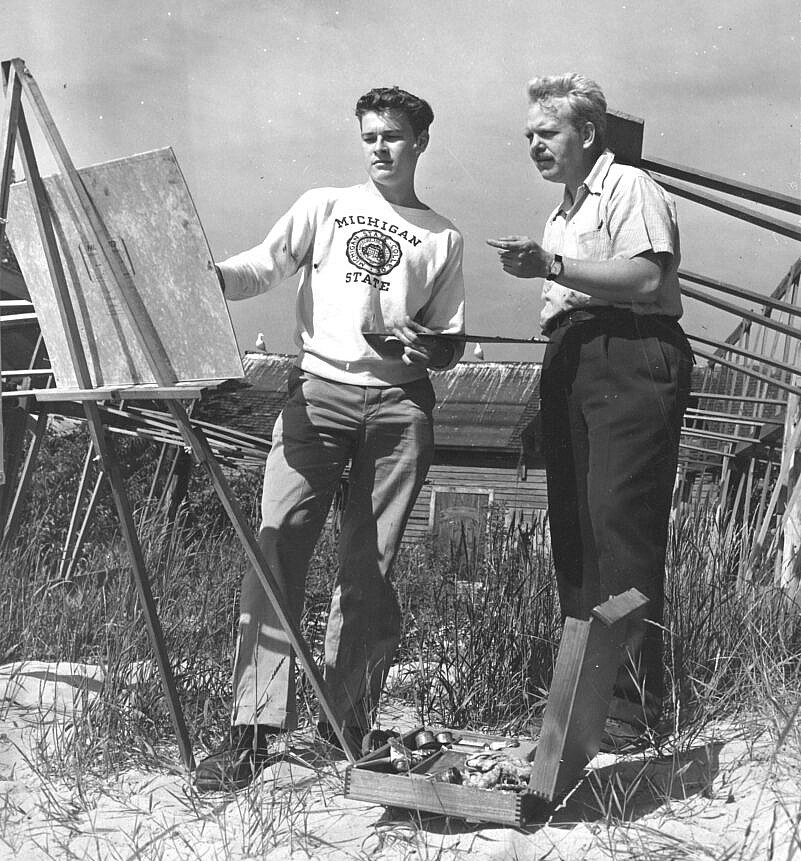 Two men standing on the beach are looking at an easel 