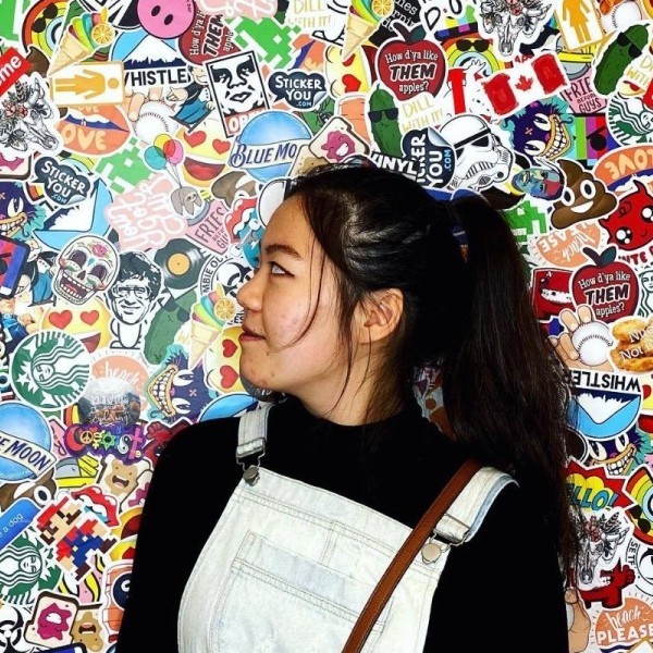 Woman with black hair in a ponytail is wearing denim overalls and a black shirt. She is standing in front of a wall full of stickers and her face is turned to the side.