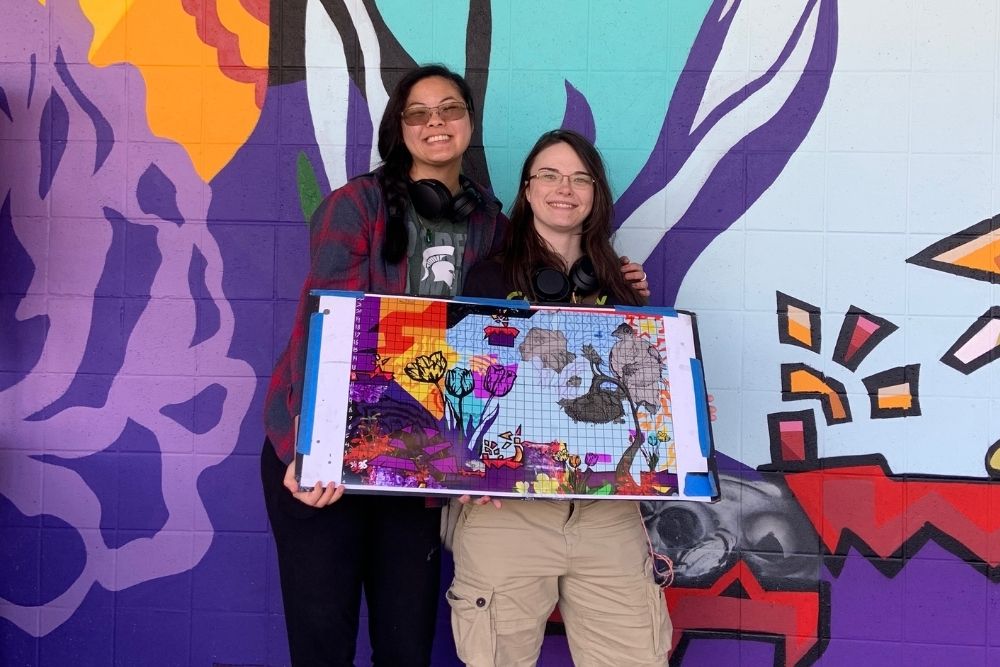 Two smiling, young women pose with a colorful sketch in front of a mural.