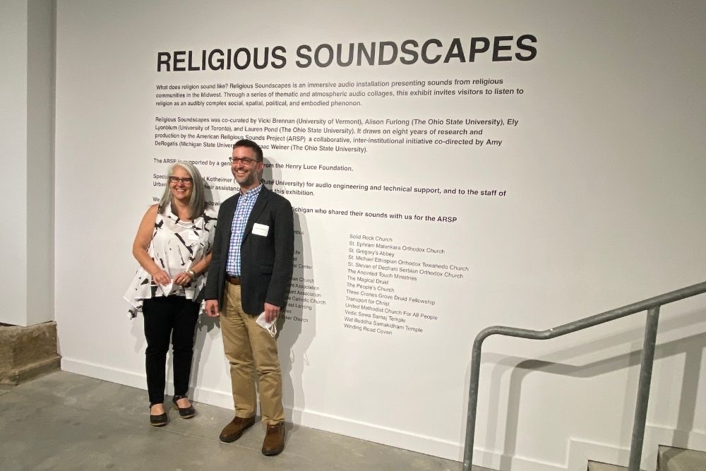 A smiling man and woman stand side-by-side in front an exhibit description printed on a white wall.