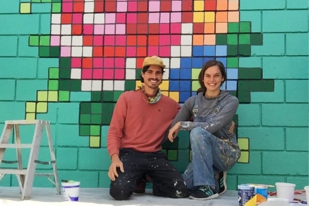 A smiling man and woman with kneel down in front of a colorful mural while surrounded by paint cans.