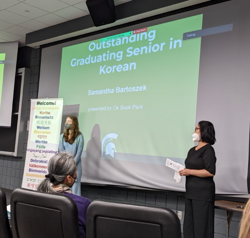 Outstanding Graduating Senior in Korean Samantha Bartoszek stands beside Ok Sook Park in front of a projected screen listing the student's name