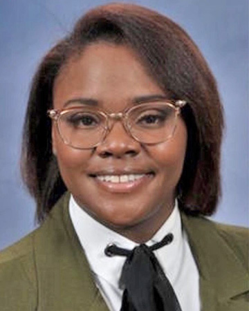 Woman with brown hair wearing glasses, a green blazer, and white undershirt with a black necktie.