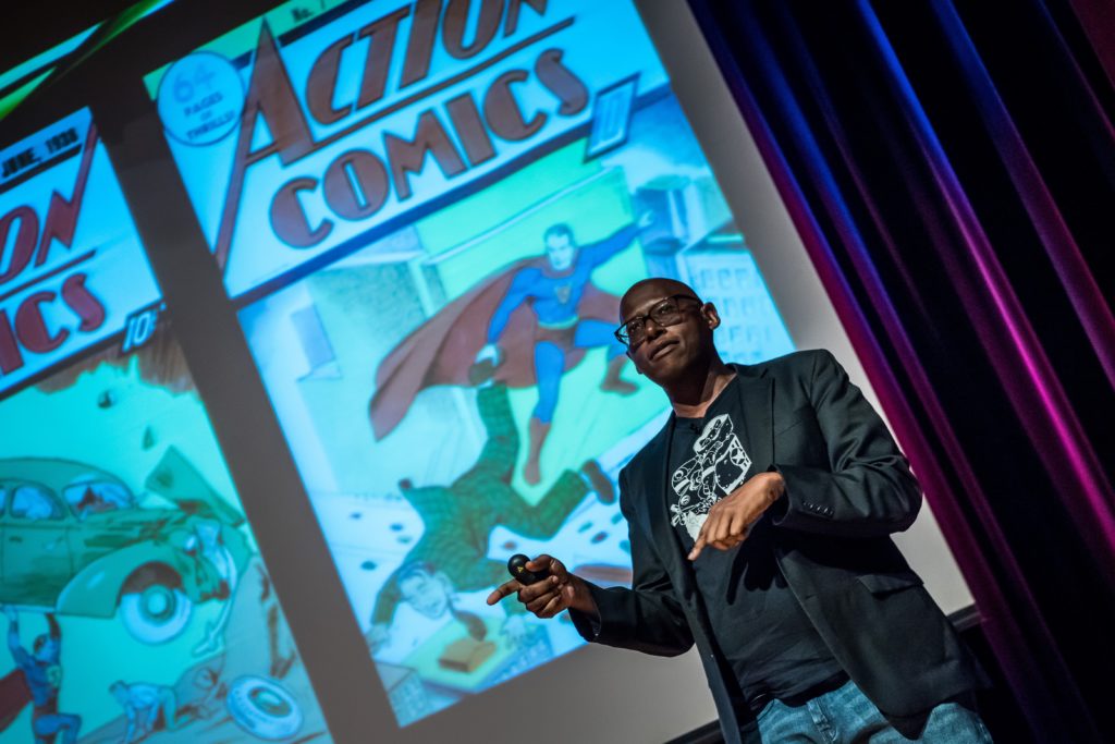 An African American man stands in front of a screen that displays covers of "Action Comics" books.