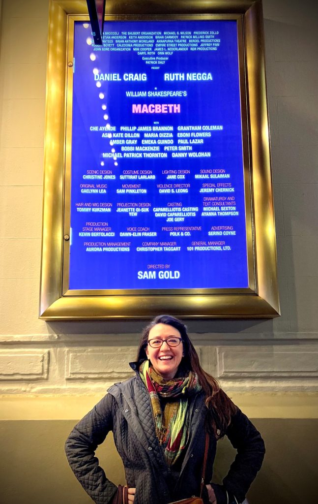 A medium shot of a smiling woman with glasses posing in front of a Broadway poster of MacBeth overhead.