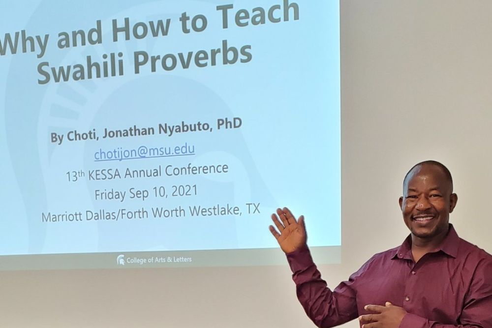 A tight medium shot of a smiling African American man gesturing towards the projected screen that reads, "Why and How to Teach Swahili Proverbs."
