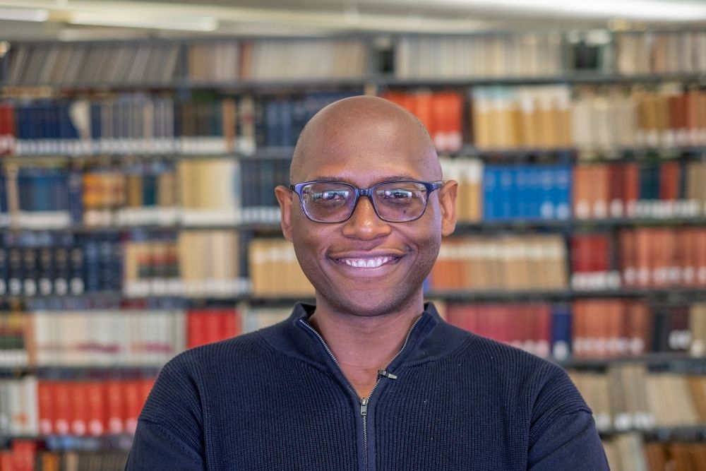 Portrait of a smiling African American man with glasses in front of a bookcase.