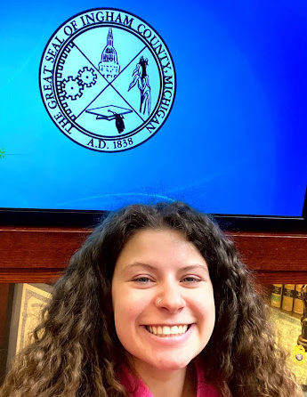 A woman smiling in front of the Ingham County Court seal.