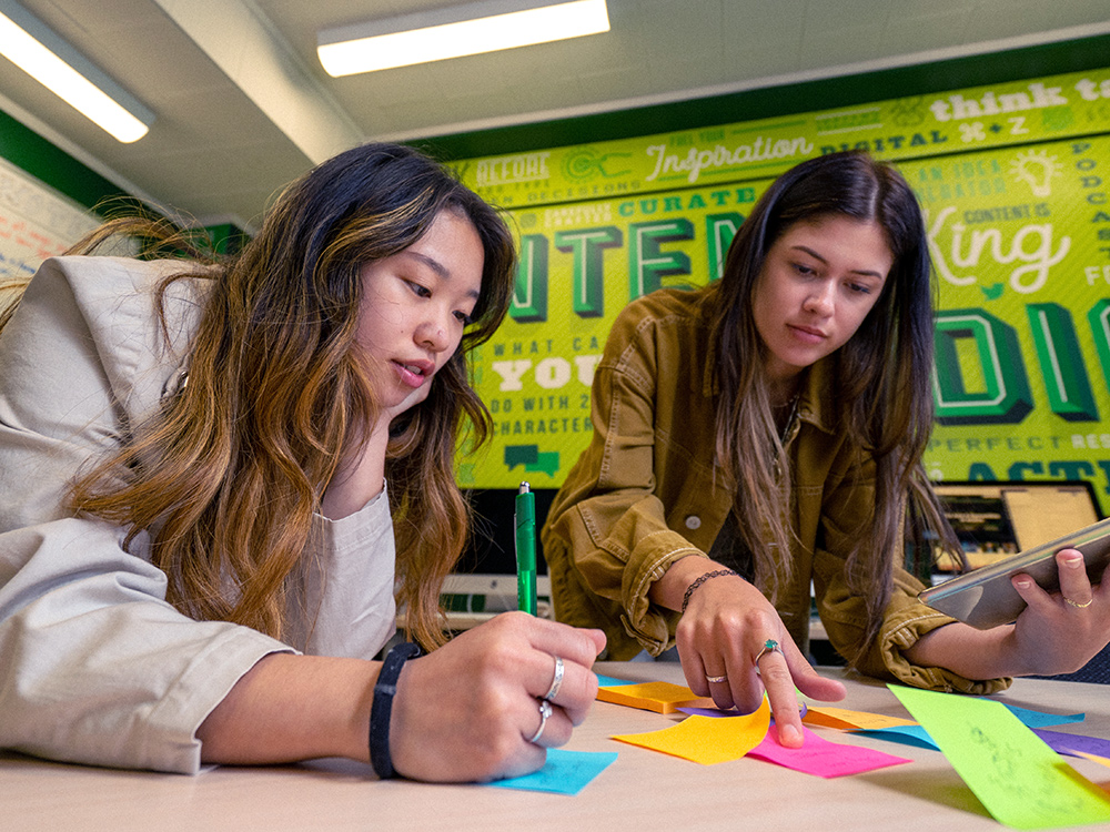 Two young women writing on Post-it Notes on a table in a colorful workspace.