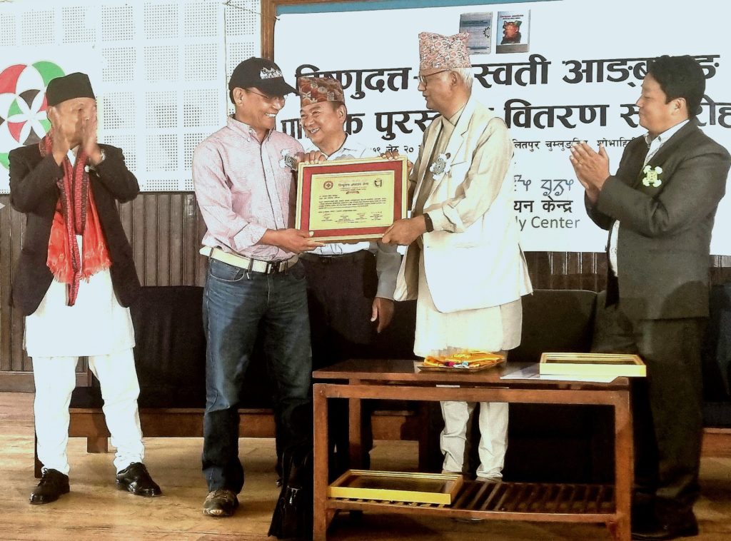 An image of a man handing an award to another man with three other men clapping in the background, indoors.