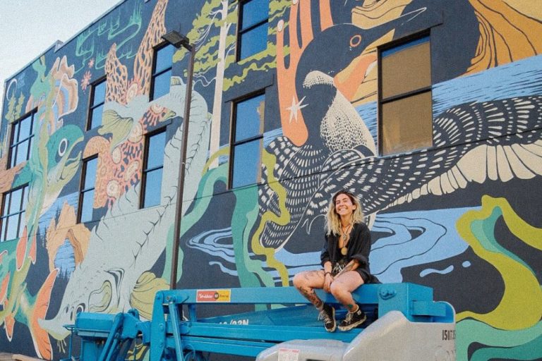 A woman sits on top of a machine in front of her mural.