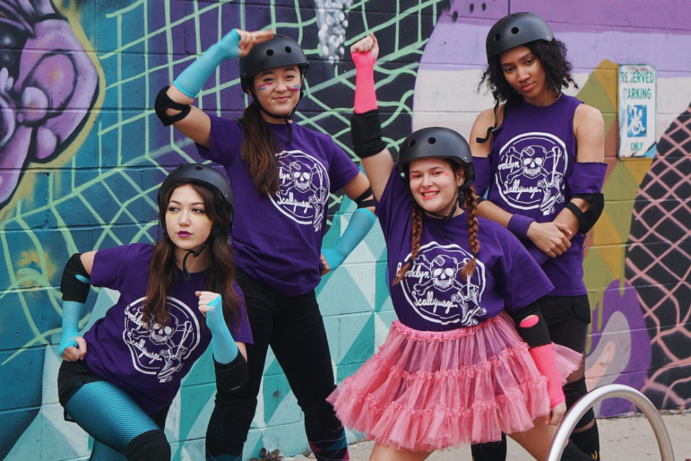 Medium shot of four young women in black helmets, purple shirts, and elbow and knee pads posing in front of a mural.