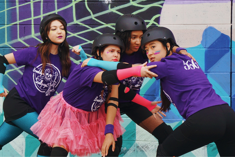Medium shot of four young women in black helmets, purple shirts, and elbow and knee pads huddling in front of a mural.