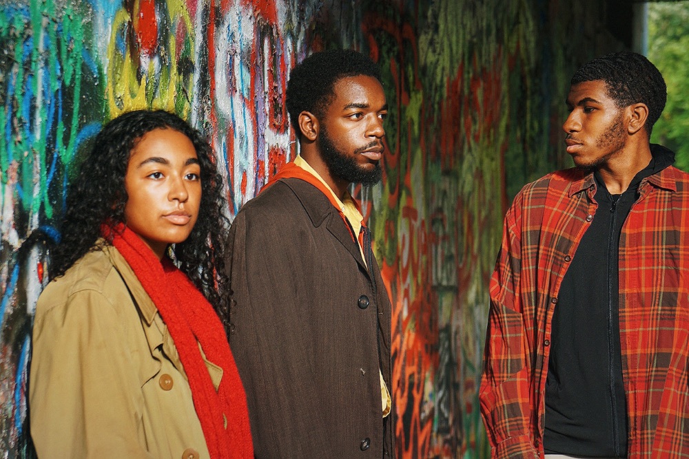 Medium shot of a young woman with dark curly hair wearing a tan coat and red scarf on the left, a young man with dark hair and beard wearing a brown coat in the center, and a young man with dark hair wearing a red plaid jacket on the right. They stand in front of a wall of graffiti. 