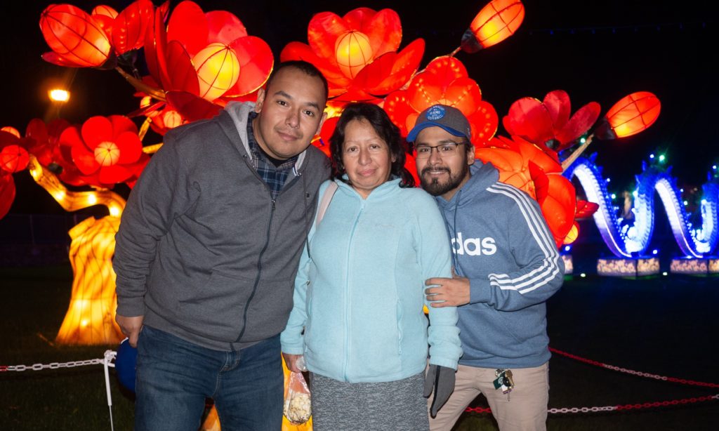 Medium full shot of two smiling men standing on both sides of a smiling woman at night outdoors. Large red decorative floral lights are overhead. 