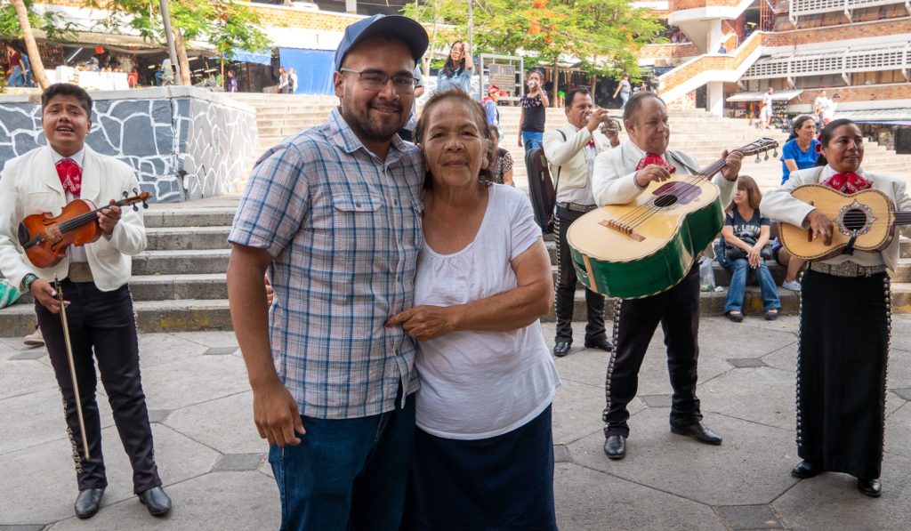 Medium shot of a smiling man with black eyeglasses wearing a baseball cap and blue plaid button-down shirt hugging a smiling older woman wearing a white shirt and black skirt outdoors. Members of a Mariachi band and a group of pedestrians stand behind them.