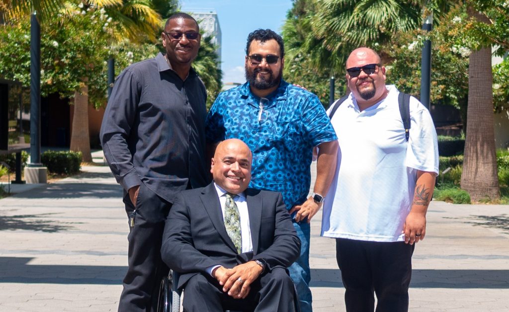 Medium full shot of three smiling men standing side-by-side behind a smiling man in a wheelchair outdoors.