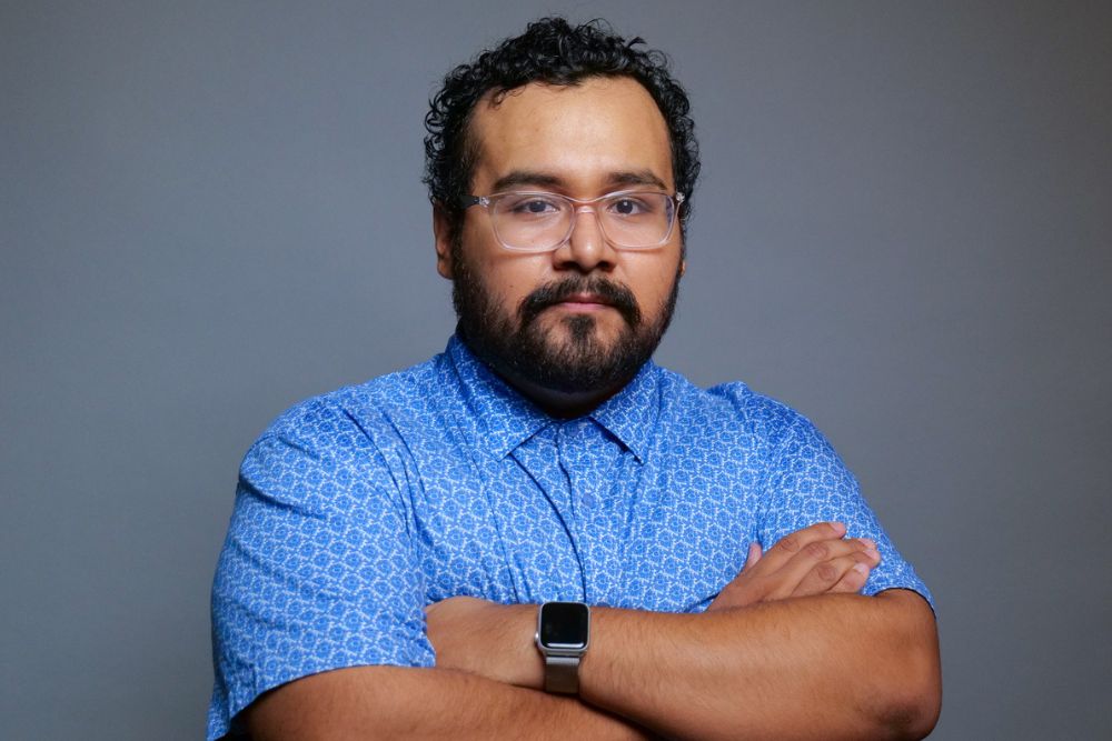 Portrait of a man with curly dark hair and a dark beard wearing clear eyeglasses and a blue printed button-down shirt.