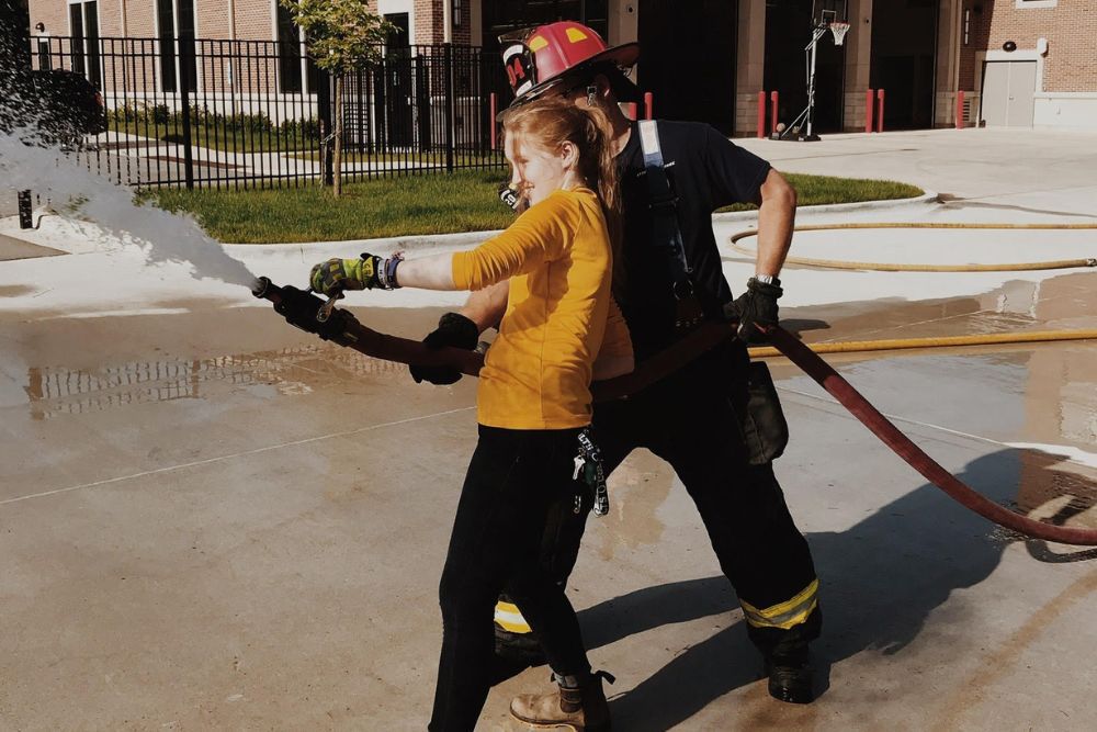 Young woman wearing a yellow shirt holds a fire hose as a firefighter stands close behind in support.