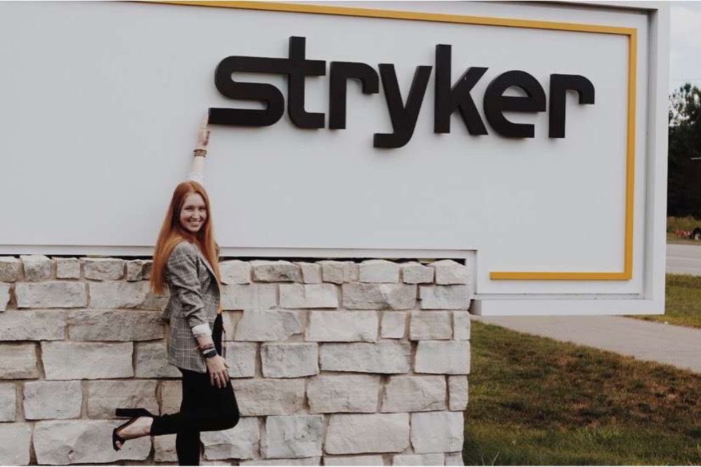Smiling young woman with red hair wearing business casual attire poses with large sign.