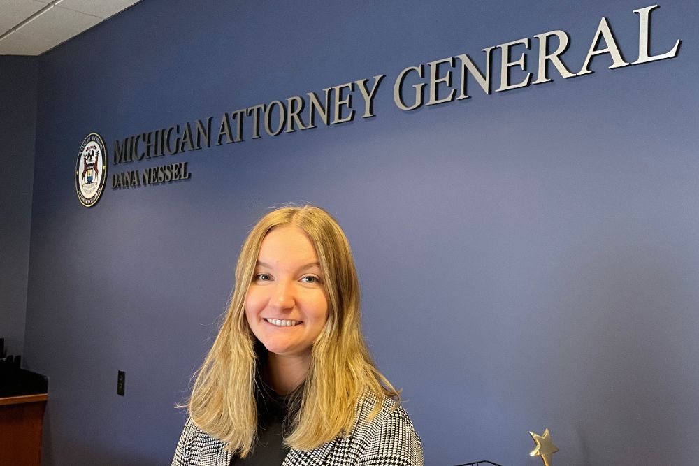 Blonde woman standing in front of  a blue wall with a Dana Nessel Michigan Attorney General's office logo.