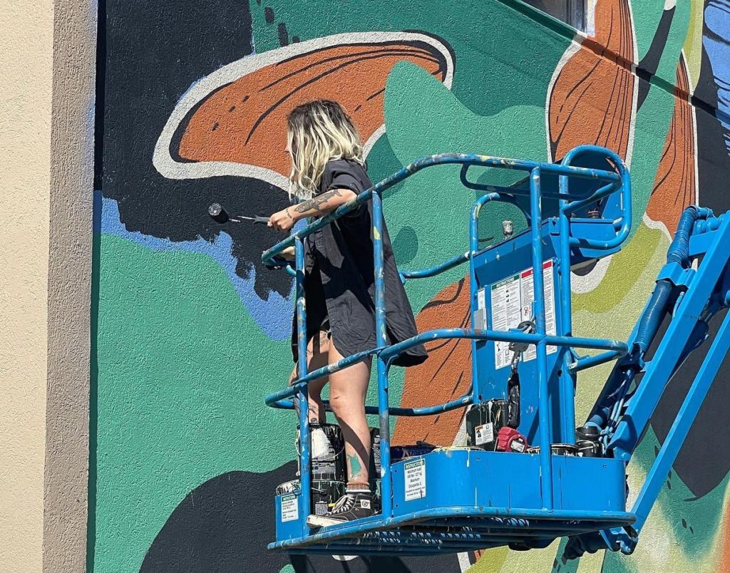 Young woman paints mural with a paint roller in hand while standing on a blue power lift machine.