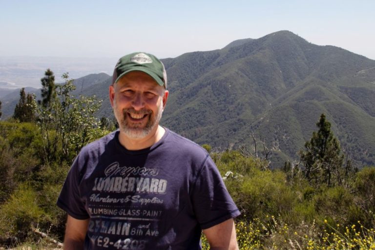 A picture of a man in a green hat and blue t-shirt smiling; there are mountains in the background.