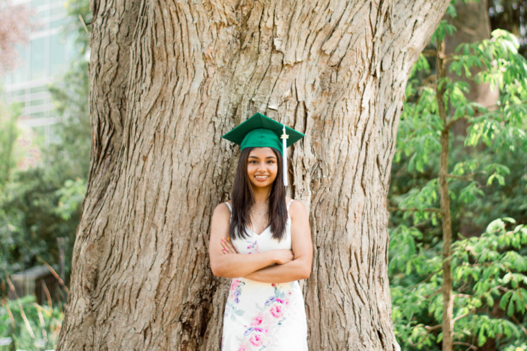 Ryda Sundilla, a recent MSU graduate, smiles and wears a graduation cap while posing in front of a tree with her arms crossed.