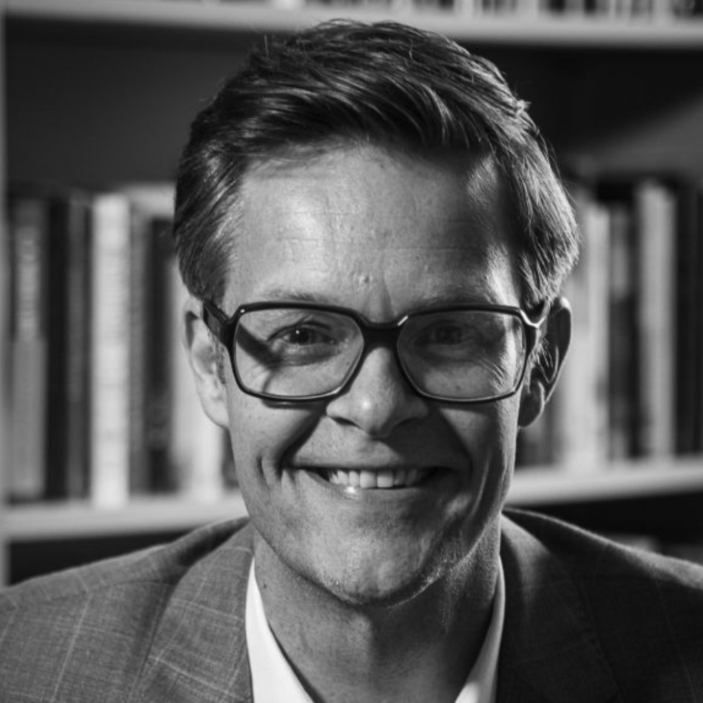 A black and white photo of a man with short hair and glasses wearing a suit smiling at the camera. 