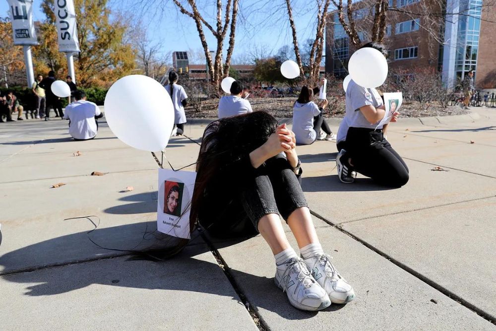 A group of people sit on cement outdoors holding white balloons and photographs of individuals.