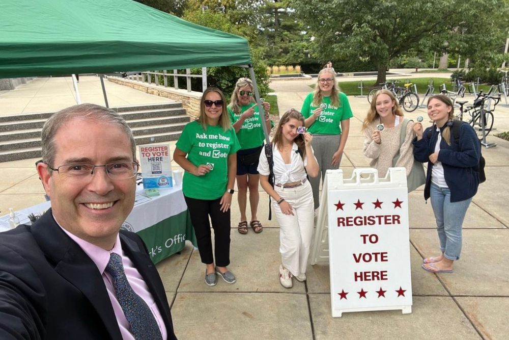 A group of smiling women stand beside a "Register to Vote Here" sign behind a man wearing glasses and a suit taking a selfie.