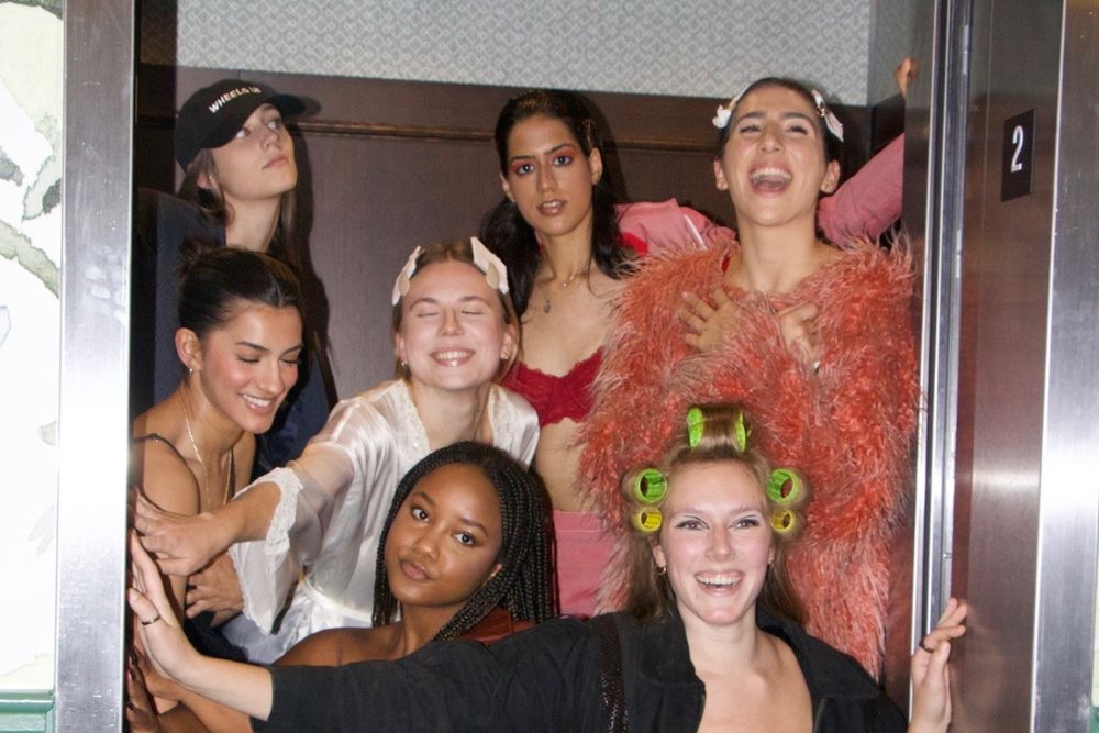 Seven young women pose inside of an elevator while holding the doors open.