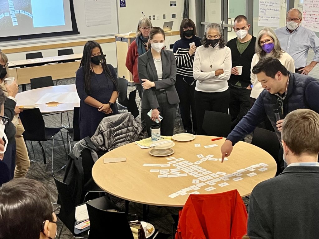 A group of people in facemasks are standing around a round table with white HuMetricsHSS values cards organized in the shape of a tree with branches and roots made of values cards. This is a paper coffee cup placed on a round paper plate at the top of the tree with another round paper plate on top of the coffee cup with a set of values cards on the plate elevated above the table. A man on the right side of the table is pointing at the roots presenting the framework.