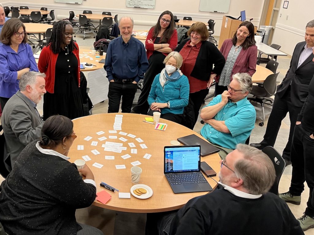 A group of people sitting around a round table with HuMetricsHSS values cards organized in the shape of a large flower - a cluster of cards in the middle of a circle with two circular rows, one inside the other. There is a column - the stem of the sunflower is a column of cards. We are looking at the flower from the top, looking down. There is a laptop on the table as well. Two rows of people stand around the table as a man in a white beard sitting at the table is presenting about the values framework.