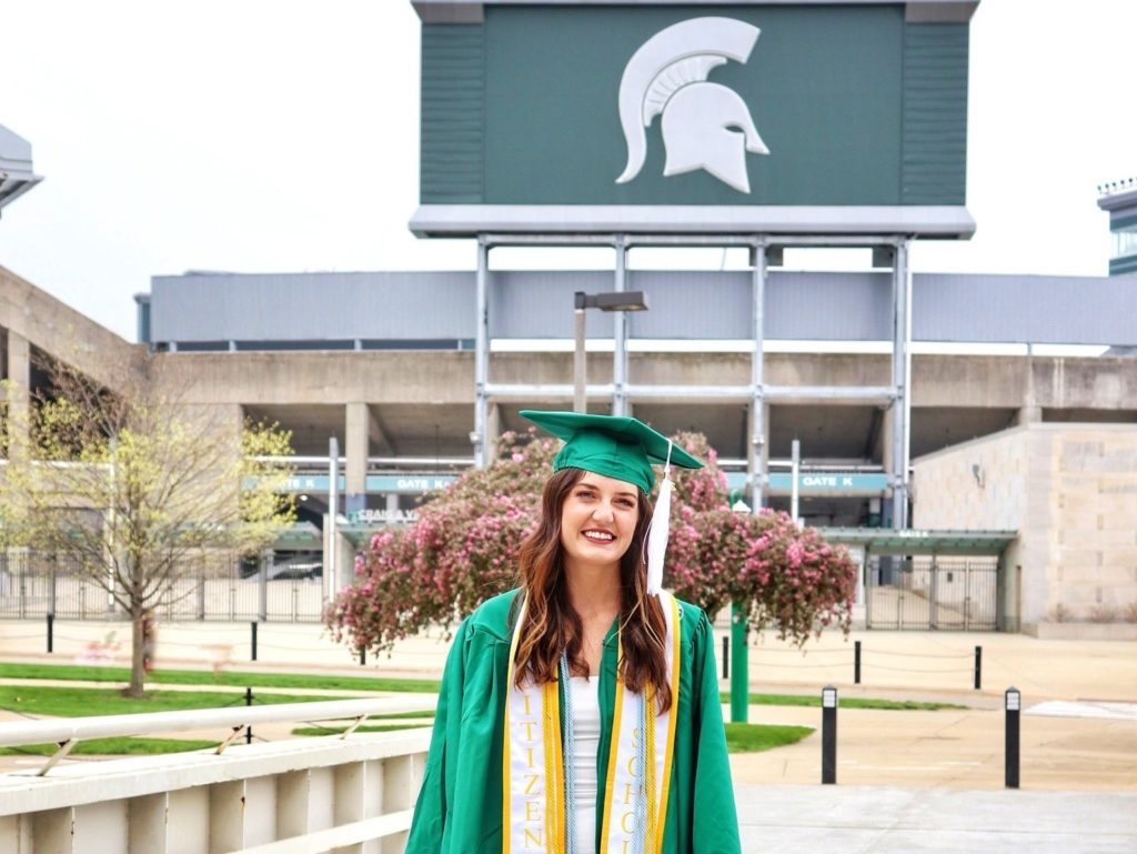 Close-up portrait of a person wearing a green graduation gown outside on a sidewalk, in front of a university logo.